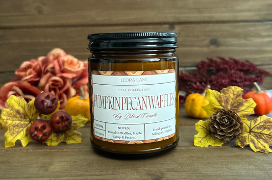 Candle jar labeled 'Pumpkin Pecan Waffles' surrounded by autumn-themed decorations on a wooden surface.