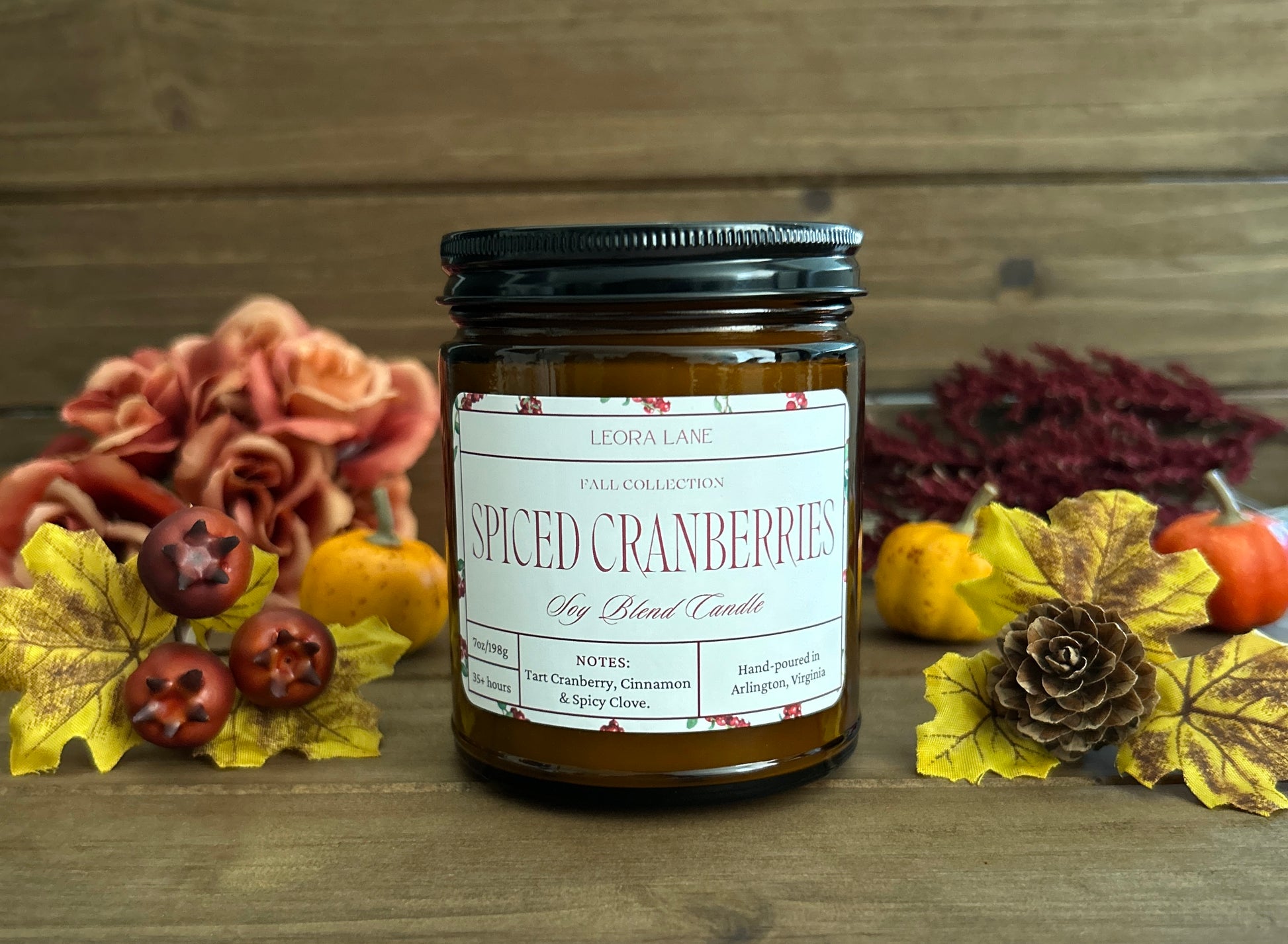 Candle jar labeled 'Spiced Cranberries' with autumn decorations on a wooden surface